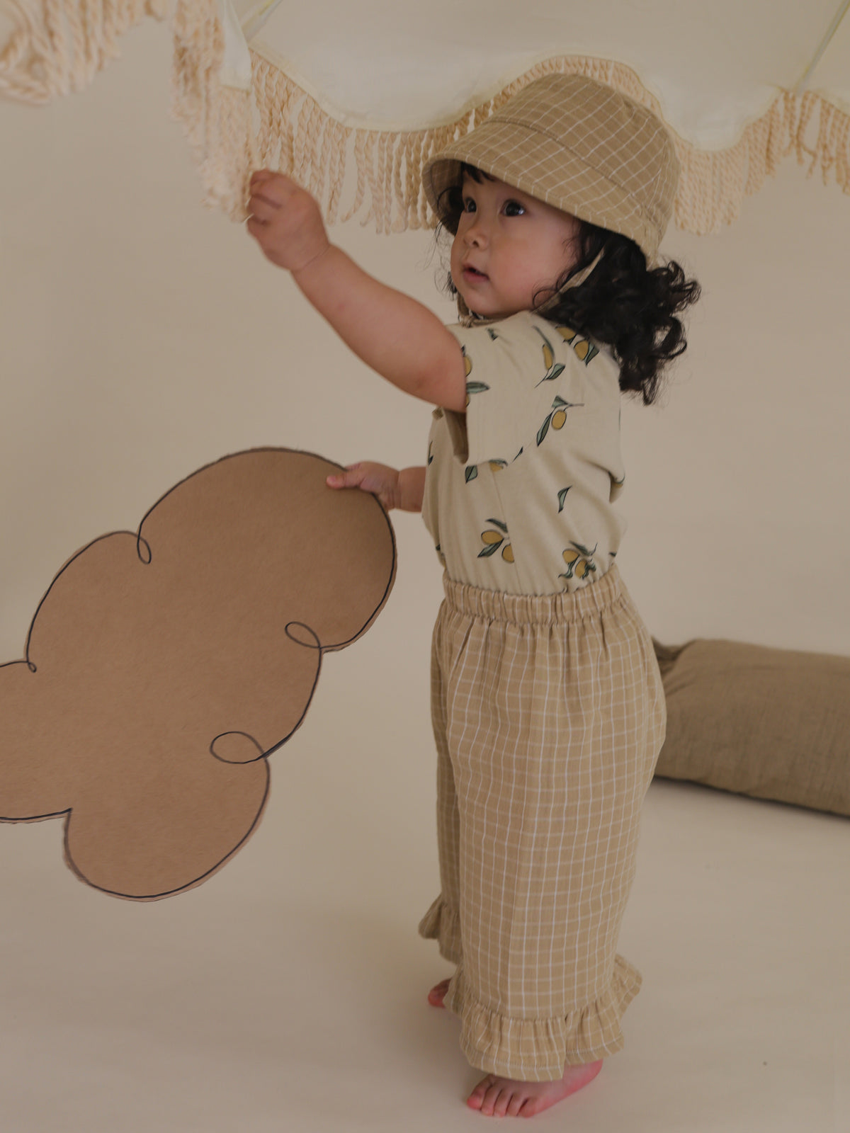 toddler wearing grid check muslin bucket hat in wheat gold with straps tied under chin pulling tassels
