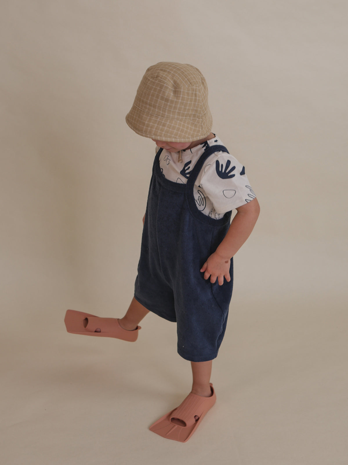 toddler in navy terry cropped overalls, t-shirt, bucket hat & swimming fins looking down standing on one leg