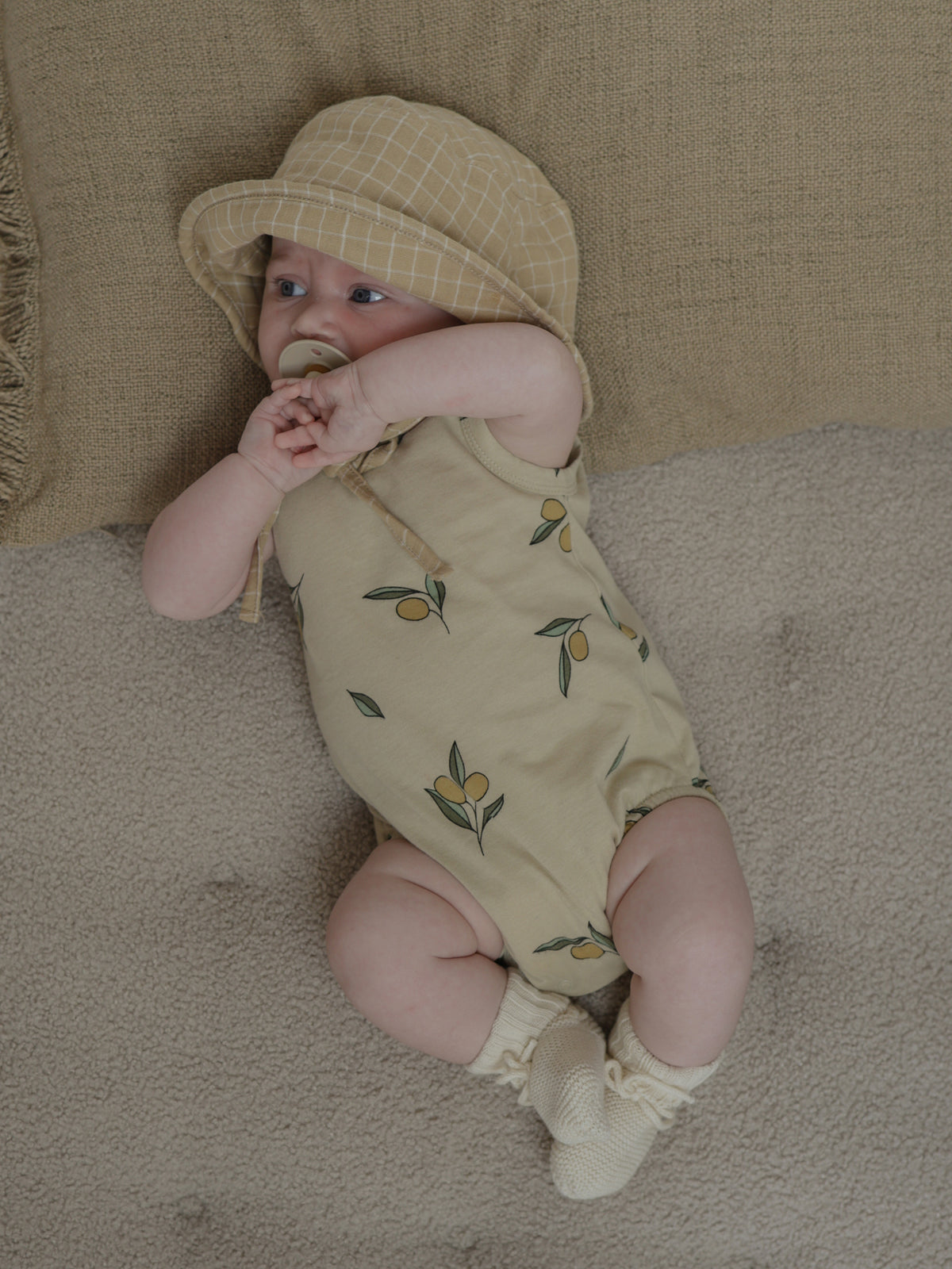 baby wearing cream sleeveless bodysuit with olives, hat & booties lying on cushion with hands together