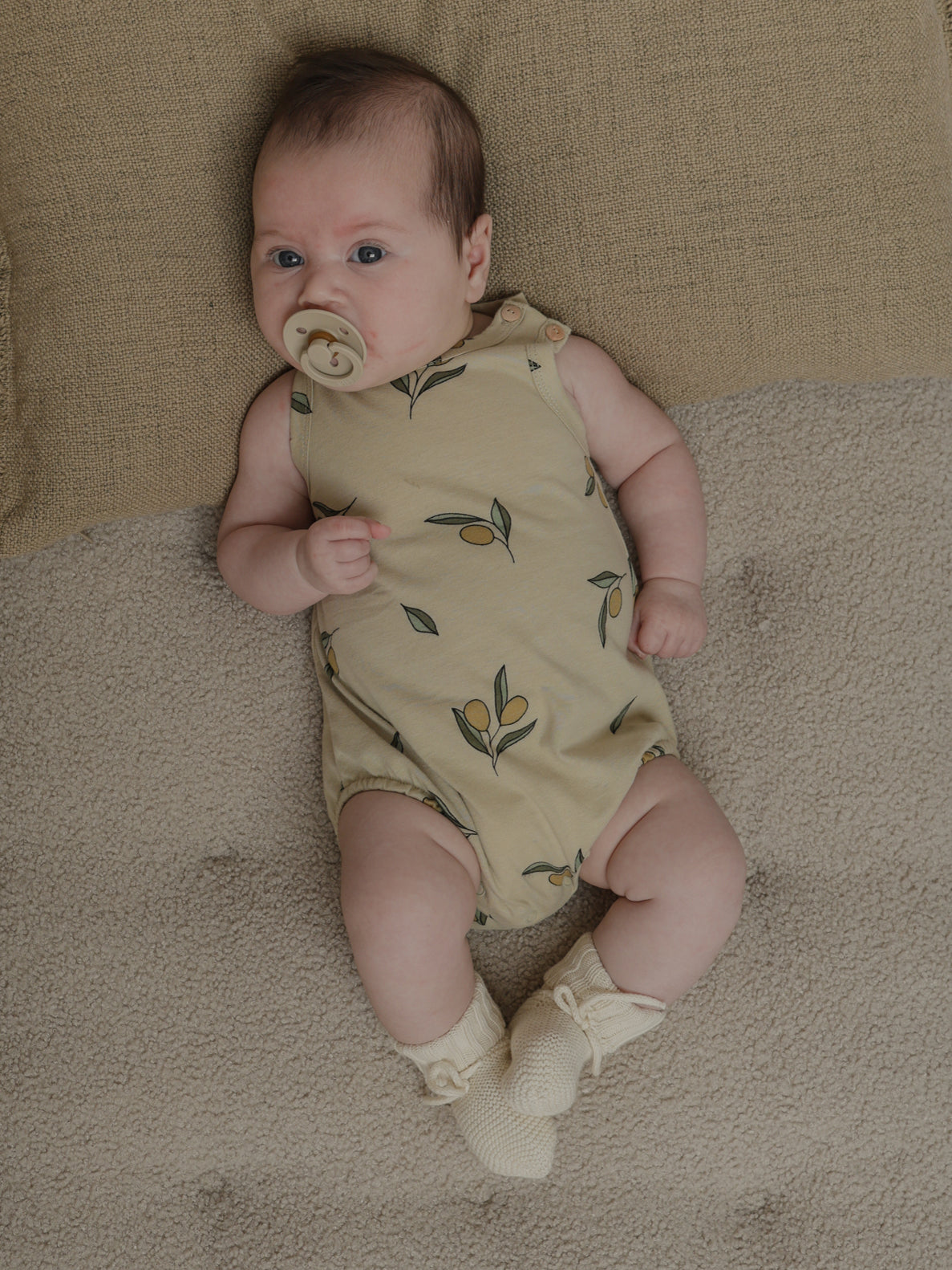 baby wearing sleeveless bodysuit with olive print & wooden shoulder buttons, with pacifier