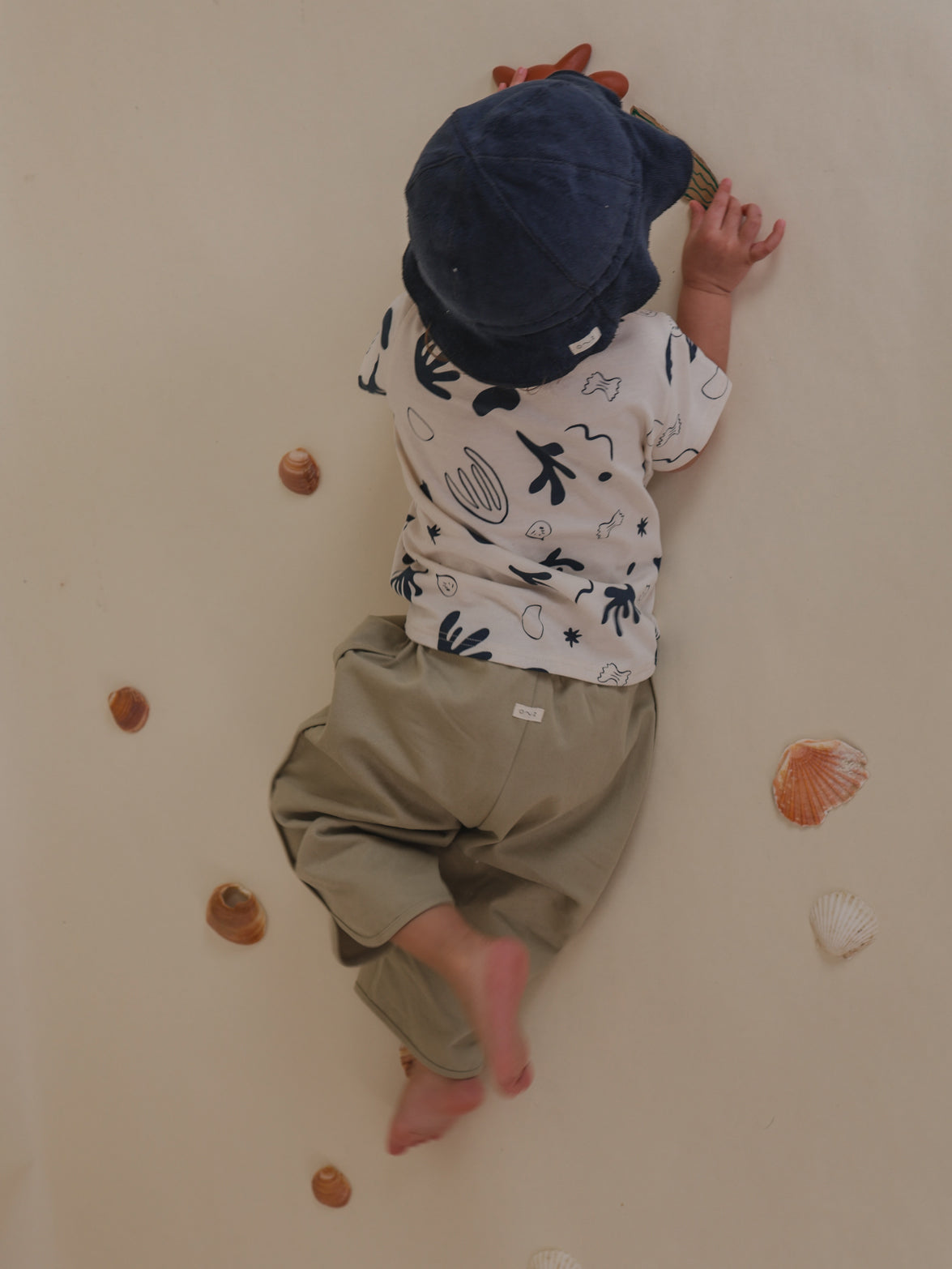 toddler in seasalt grey balloon leg pants, t-shirt & sun hat, playing on the floor with seashells
