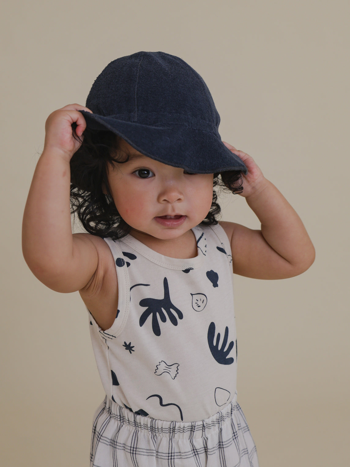 toddler playing with brim of terry sun hat in navy, wearing printed vest, grid pants