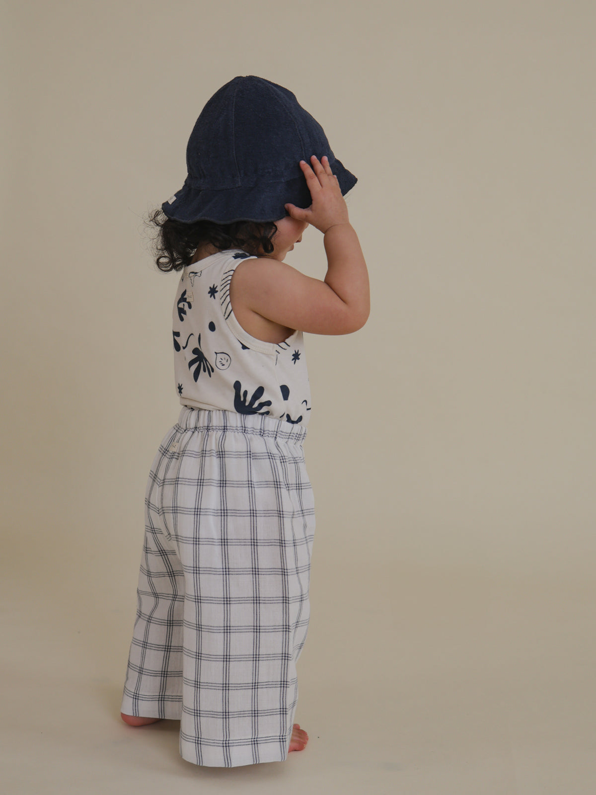 side view of toddler in navy terry sun hat, printed vest, grid trousers, holding the brim of hat