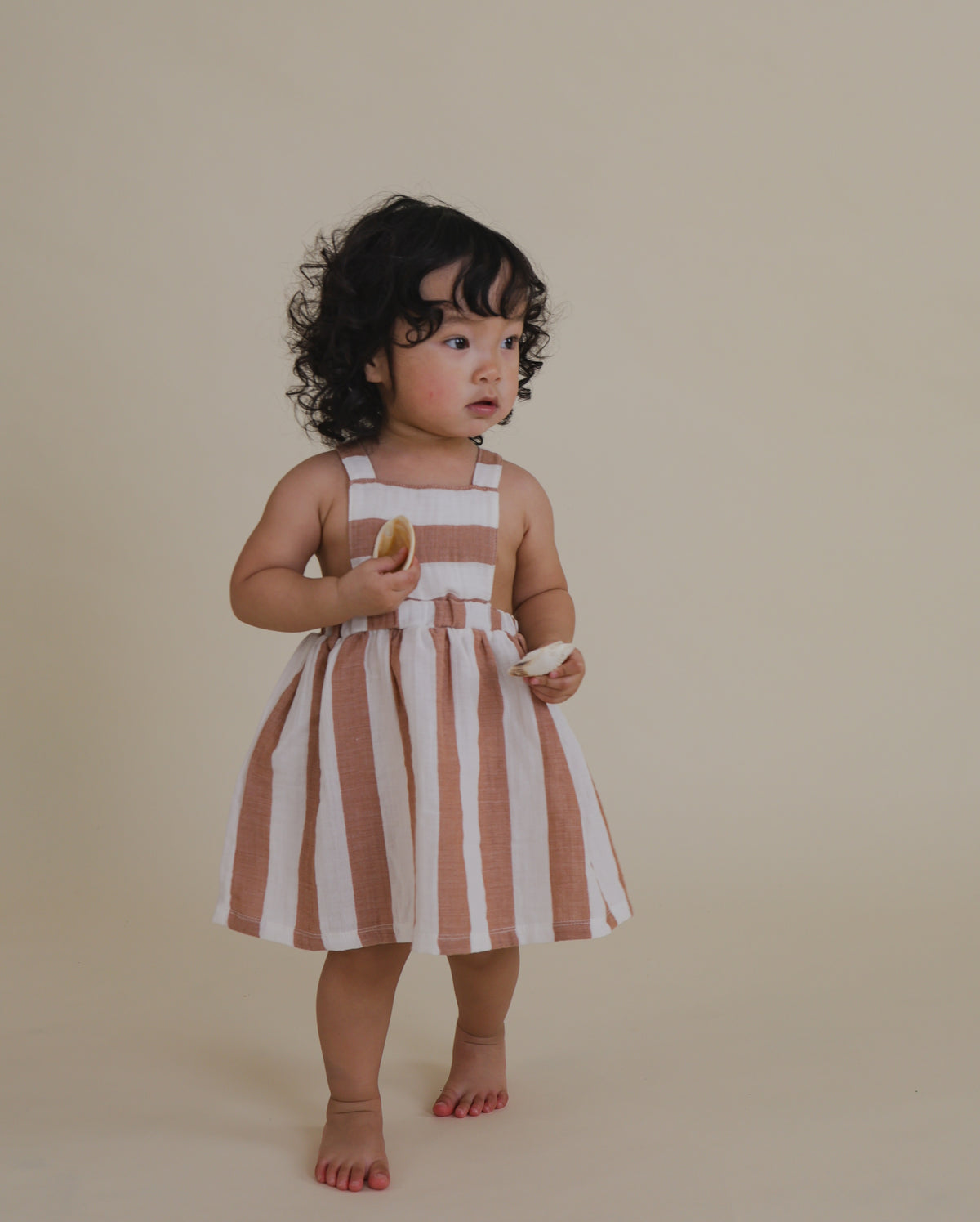 toddler wearing red striped apron skirt in soft organic muslin holding seashells