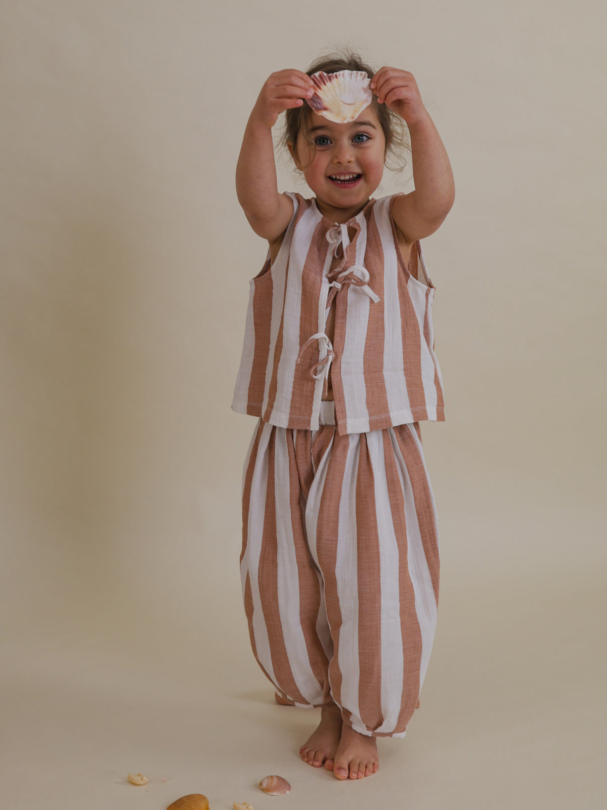 happy toddler wearing two way muslin ribbon front-tie resort top with red stripes holding seashell