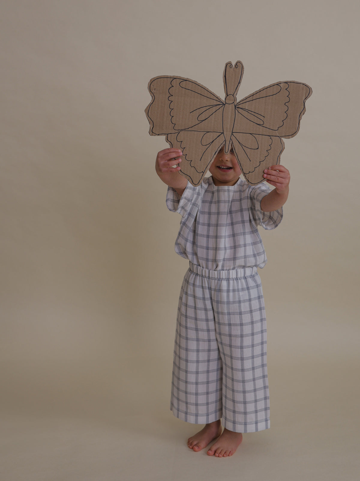 toddler in navy window check muslin boxy t-shirt tucked in, holding cardboard butterfly