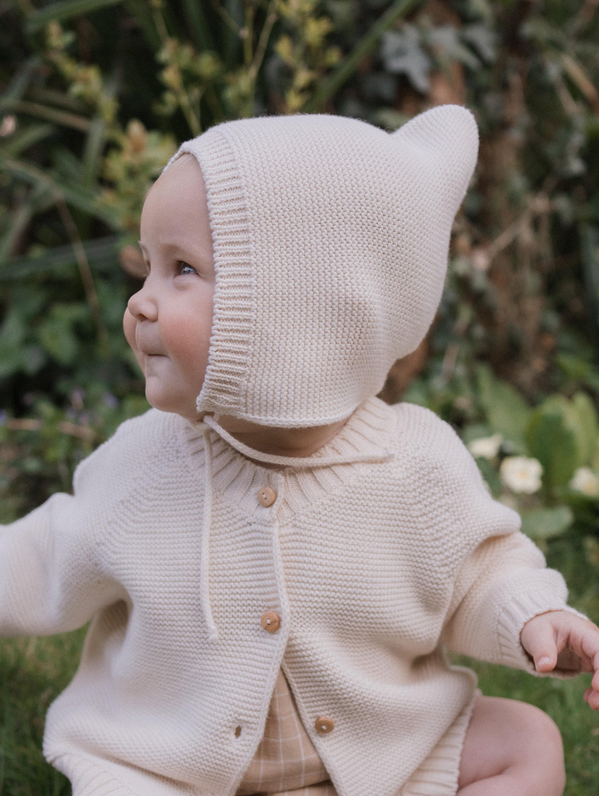 happy baby in knitted cotton pixie hat with chin tie and matching cardigan, sitting outdoors