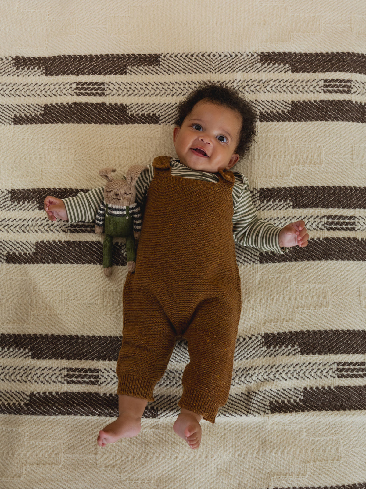 baby in chestnut brown wool salopette and striped top, with matching wool bunny toy in arms