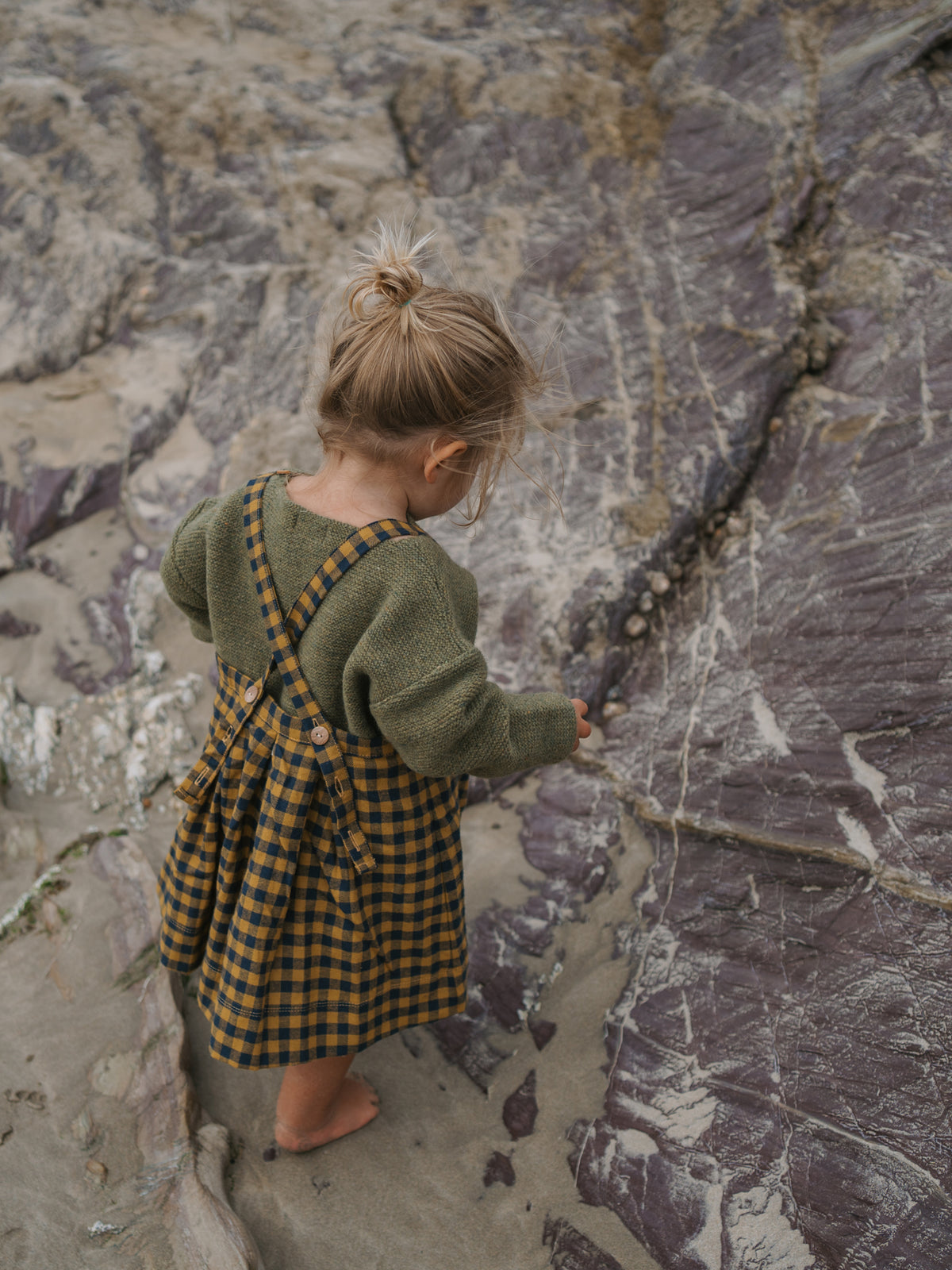 Child in a green cardigan and checkered dress standing on a rocky surface.