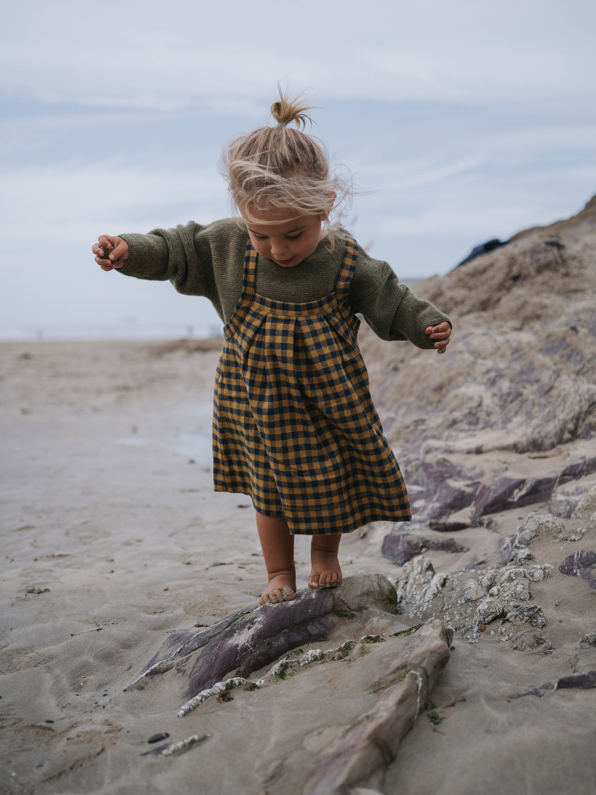 Child in a green sweater and checkered dress standing on a rocky beach.
