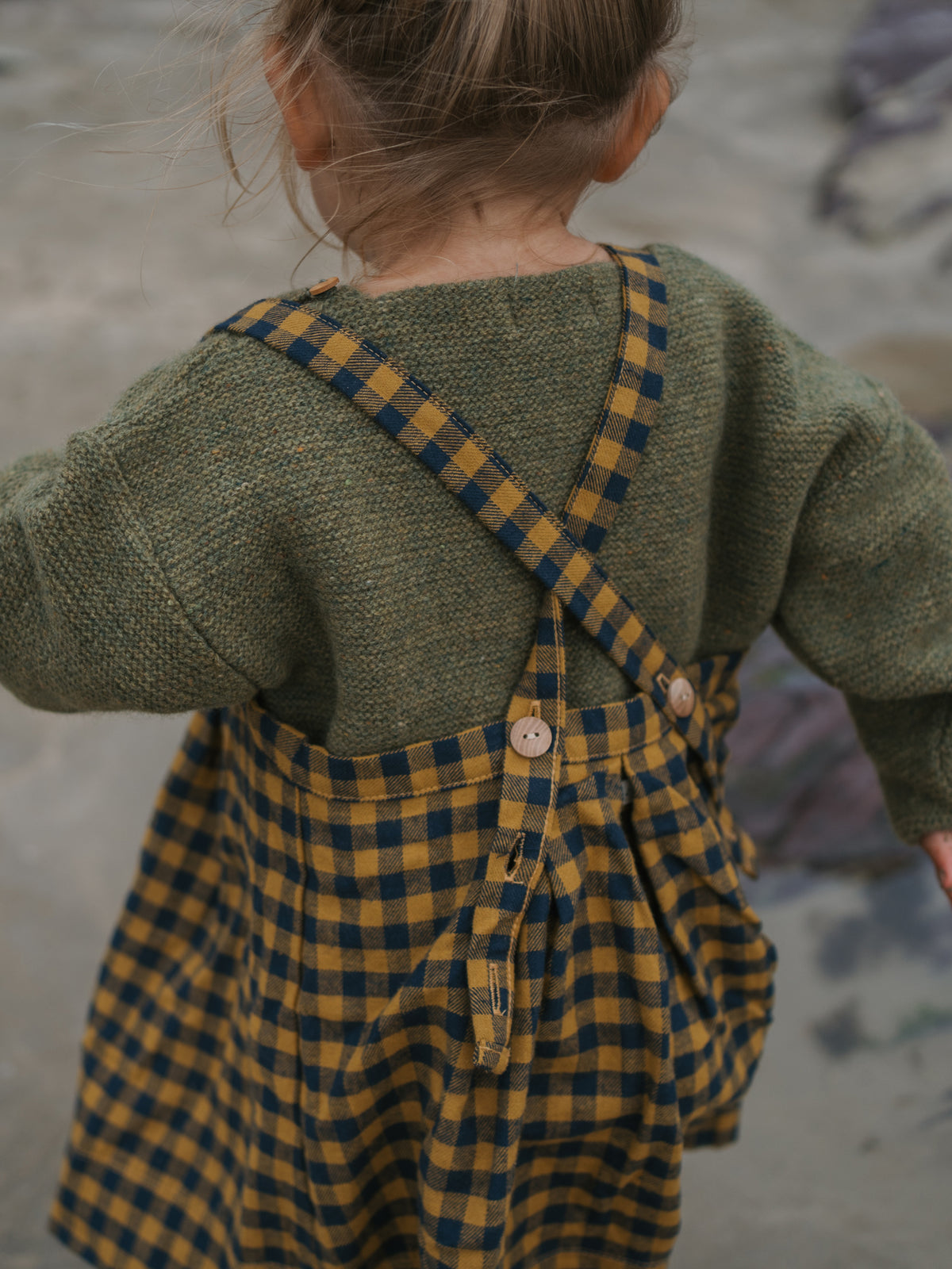 Child wearing a green sweater and yellow checkered pinafore dress on a beach.