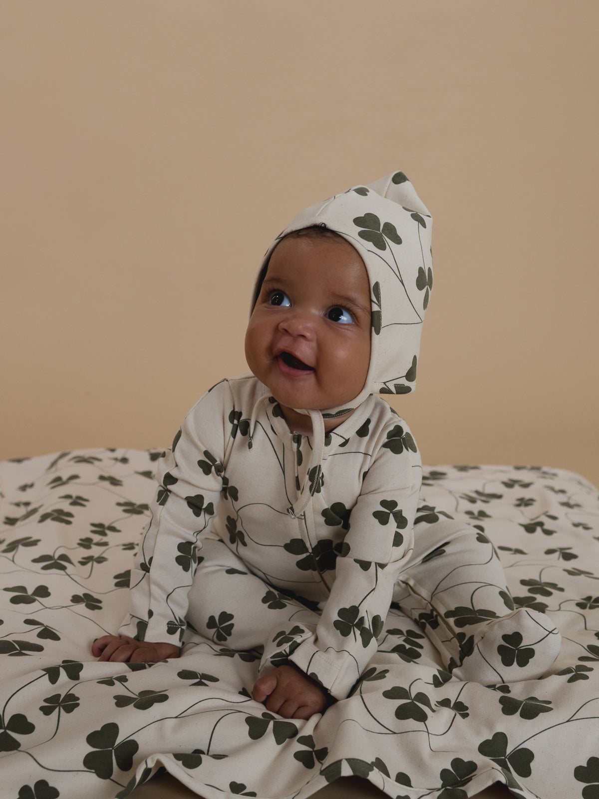 baby in matching cream pixie bonnet and onesie with green clover print, sitting on matching blanket