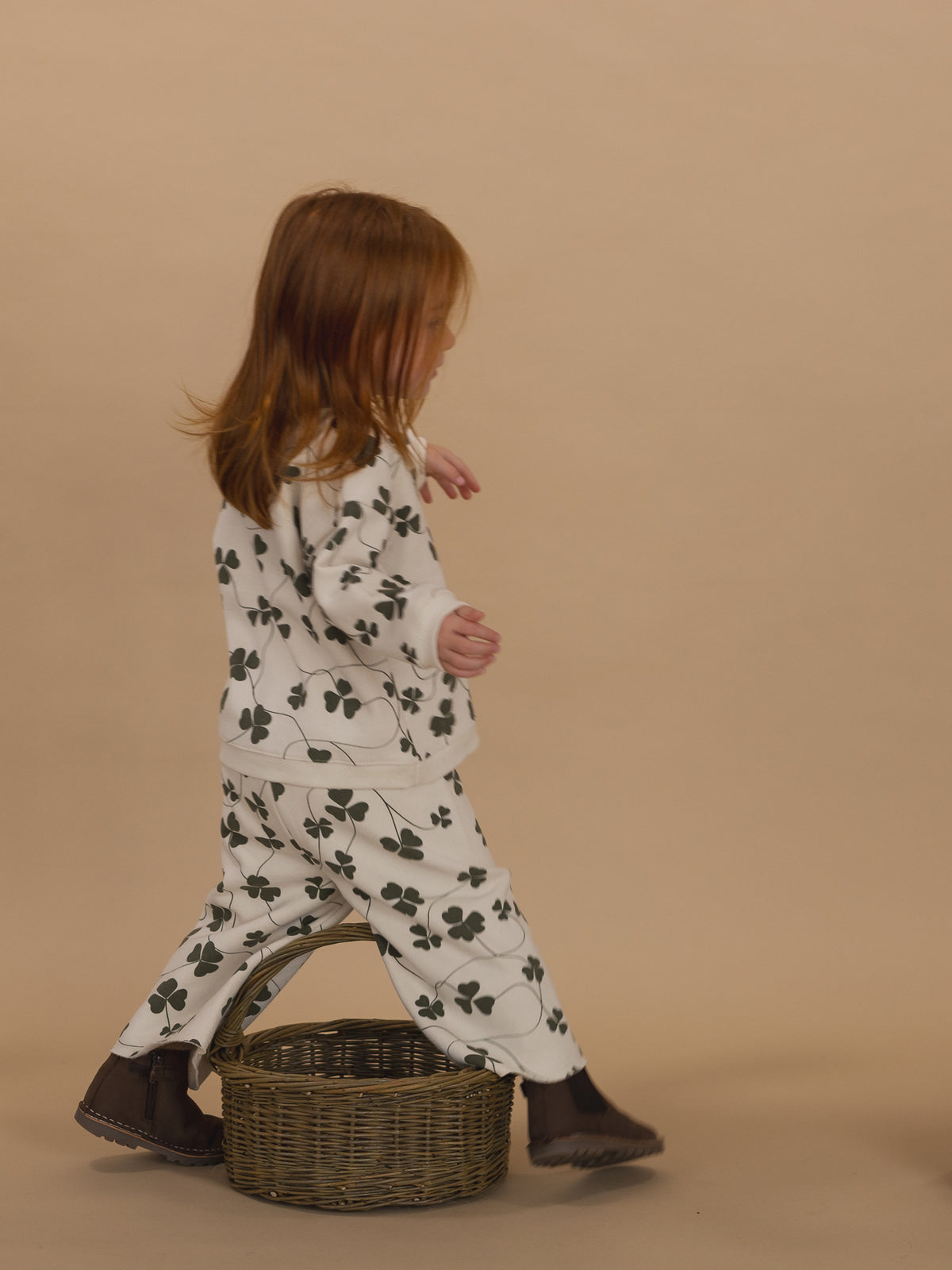 toddler in matching cream sweatshirt and pants with green clover print, crossing over rattan basket