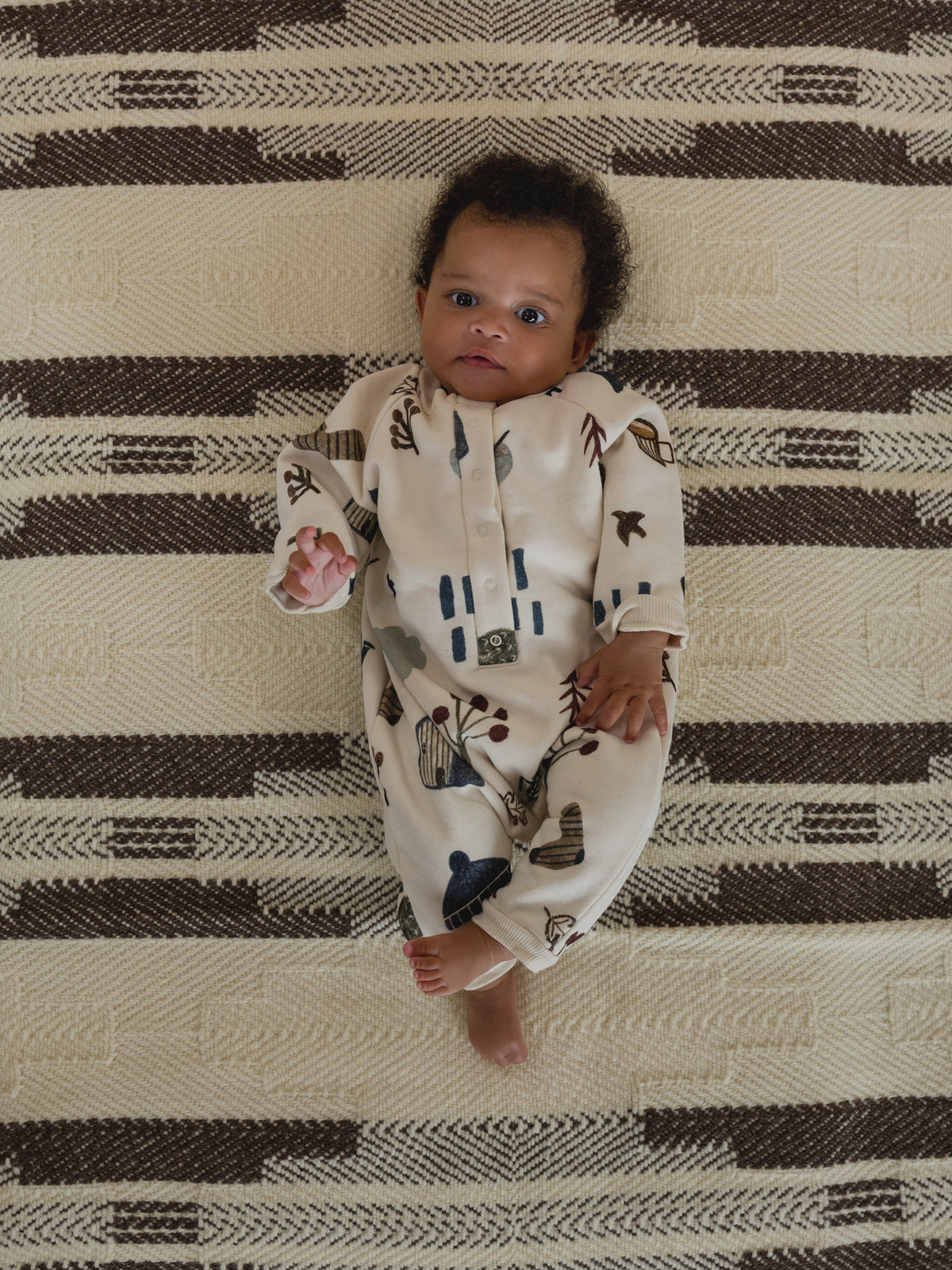 baby in cream undyed cotton onesie with cosy nature-themed hand-drawn prints, lying on carpet
