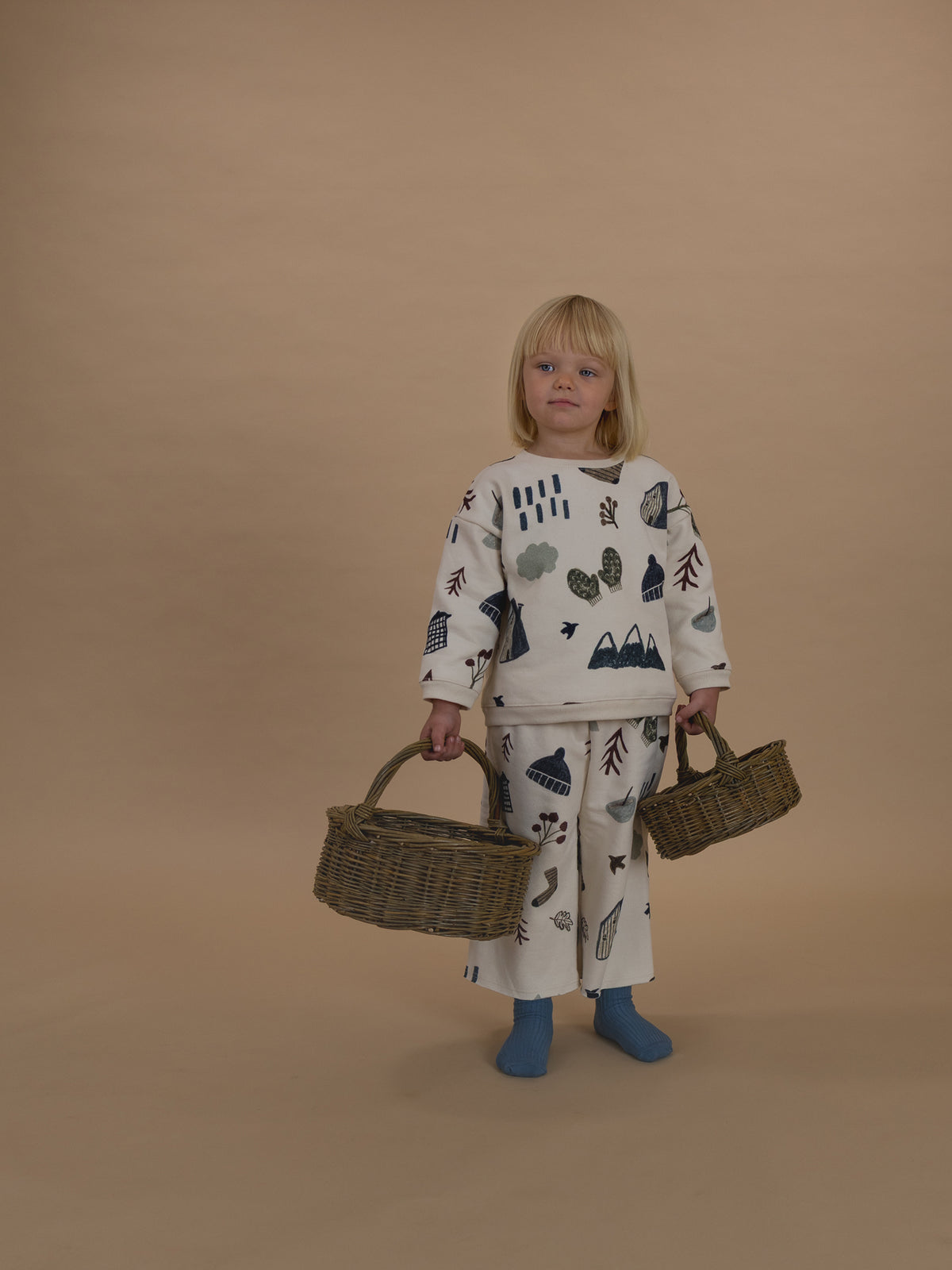 toddler in matching cream cotton sweatshirt and pants with cosy nature-themed graphics, holding baskets
