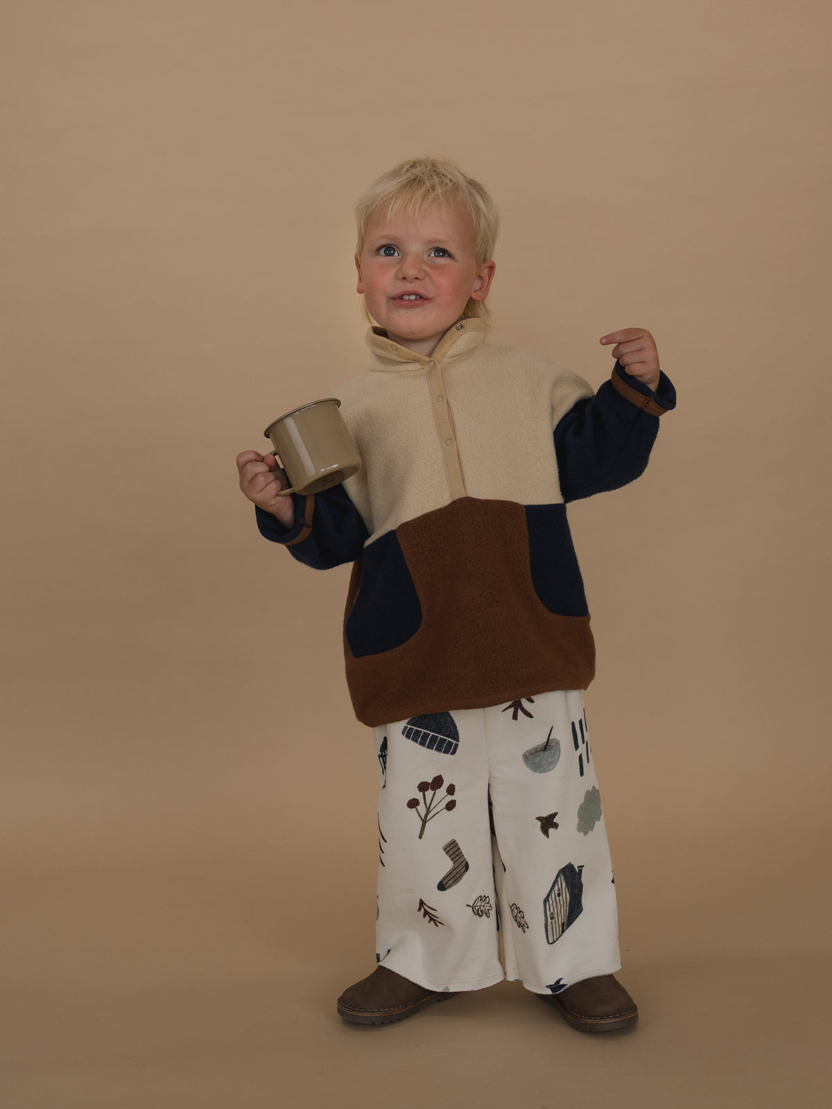 toddler in wide leg pants with cosy nature-themed print & colour block sweater, posing with a cup