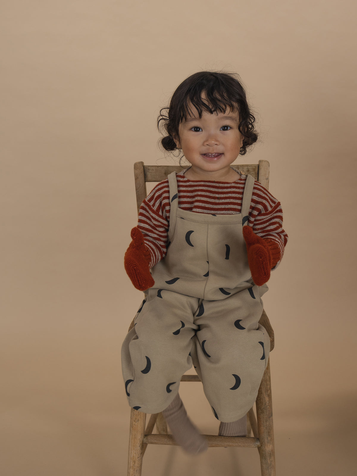 toddler in beige dungarees with black moon print, red striped sweatshirt and gloves, sitting on chair