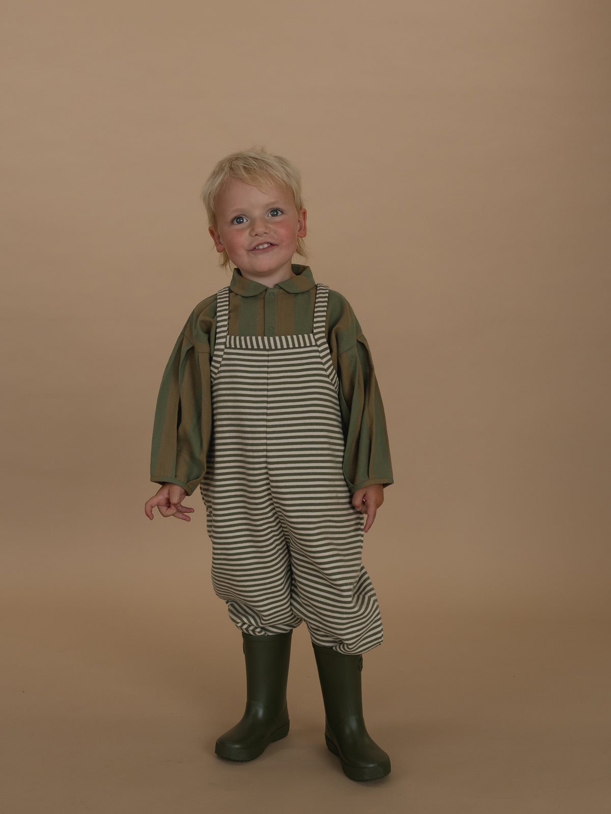 toddler in oversized boxy green and white striped dungarees with lodge shirt and rain boots