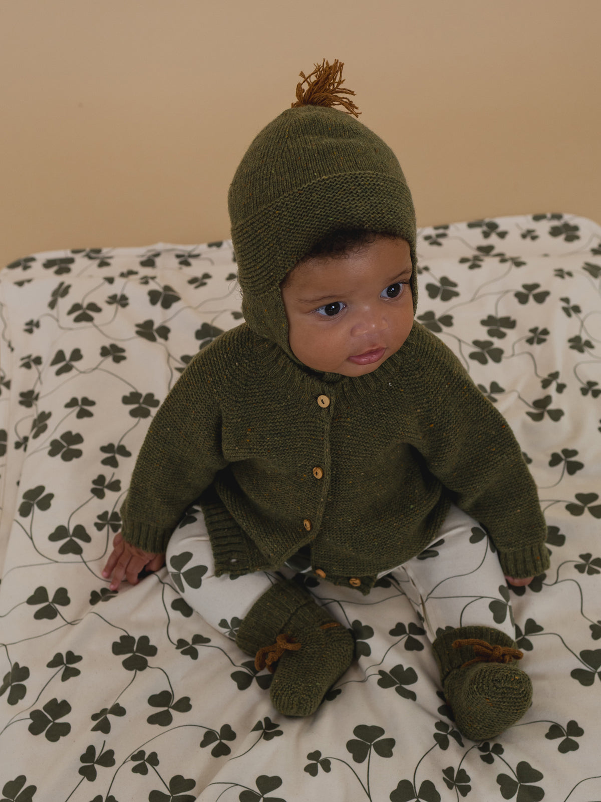 baby in forest green wool booties with brown ribbons, matching cardigan and tassel hat, clover sleepsuit