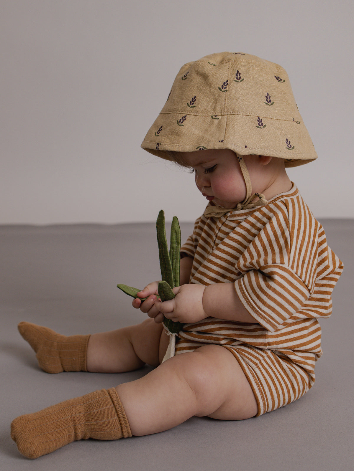 Child wearing a striped outfit and sun hat holding green beans on a plain background