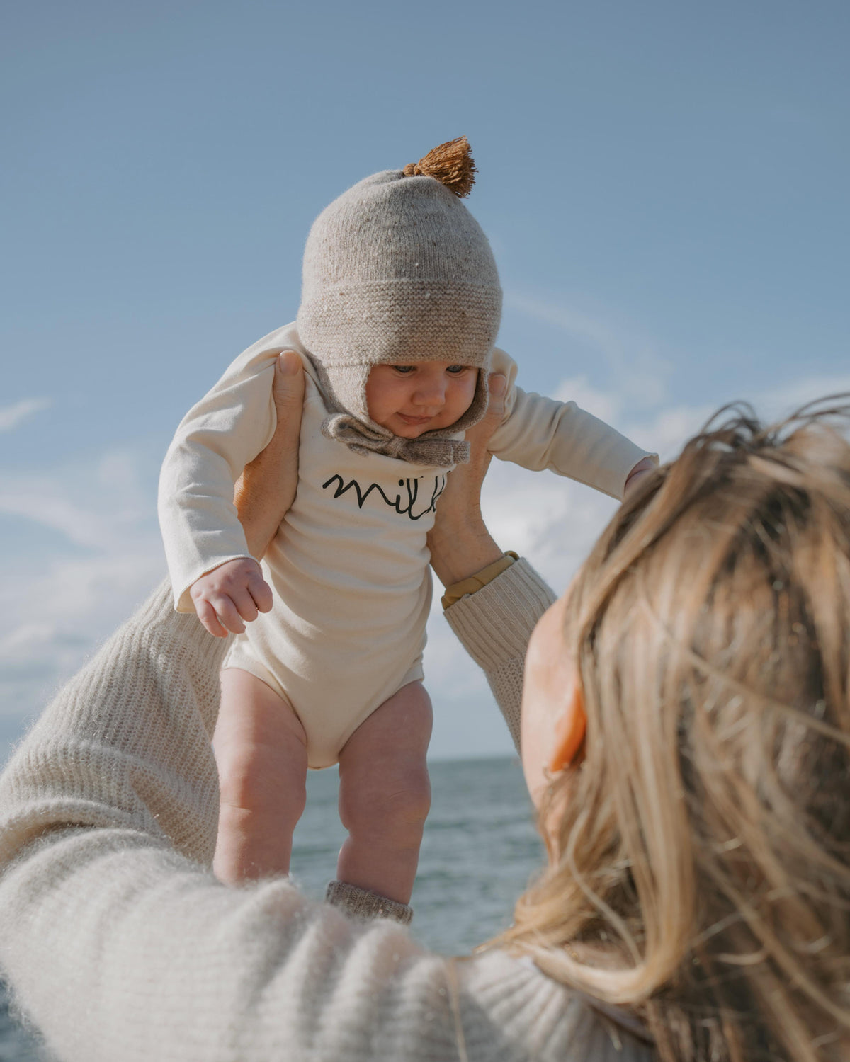 Baby in a milk undyed cotton bodysuit and hat being held by an adult on a beach.