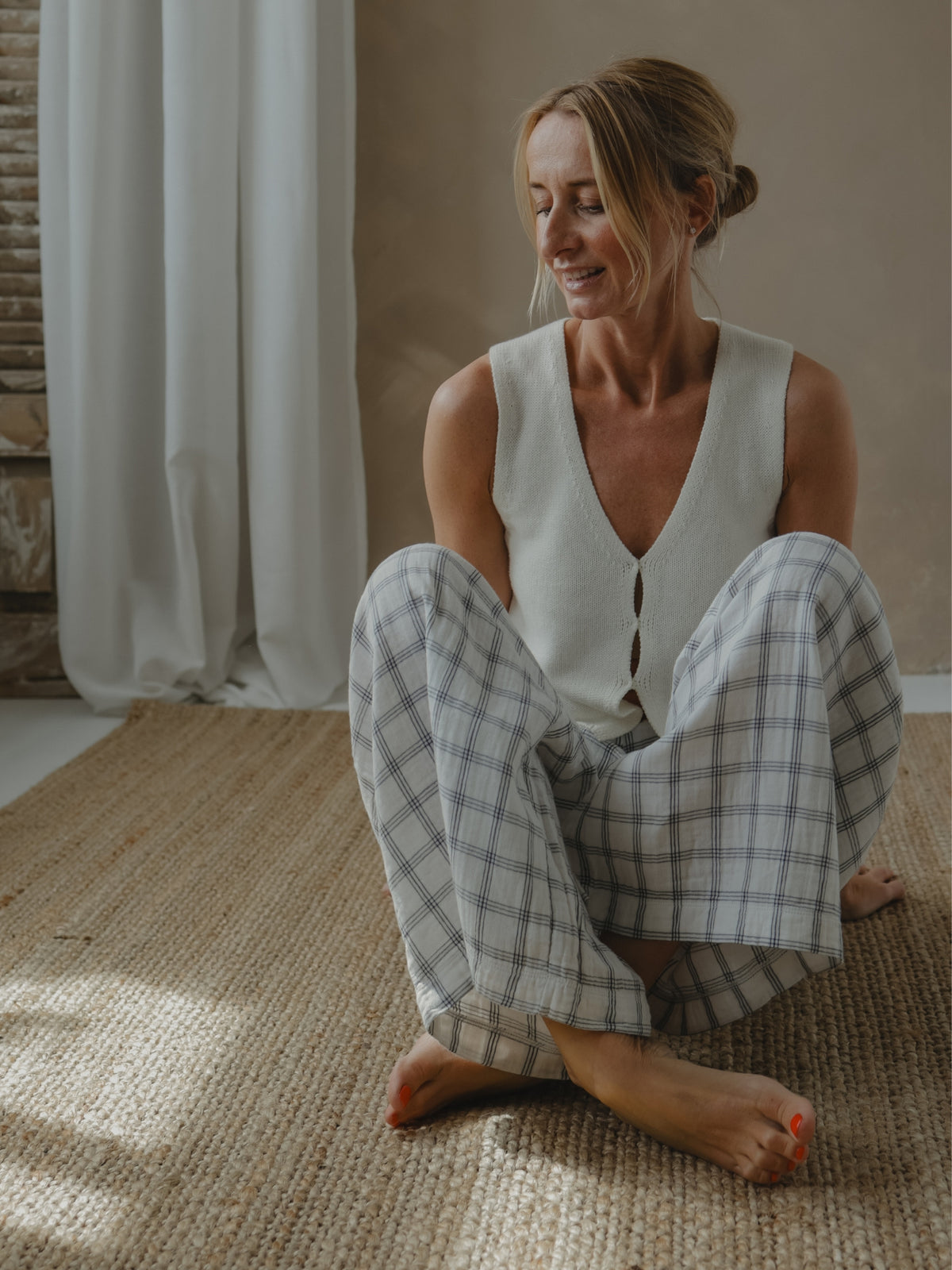 Woman sitting on a woven mat wearing a white sleeveless top and checkered pants in a softly lit room.