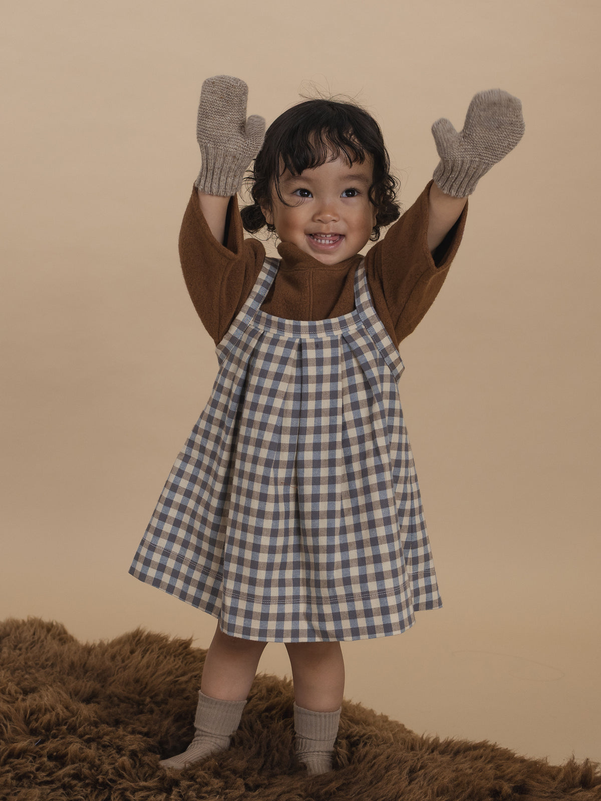 toddler in oatmeal mittens with ribbed cuffs, brown jumper and gingham skirt standing with hands up