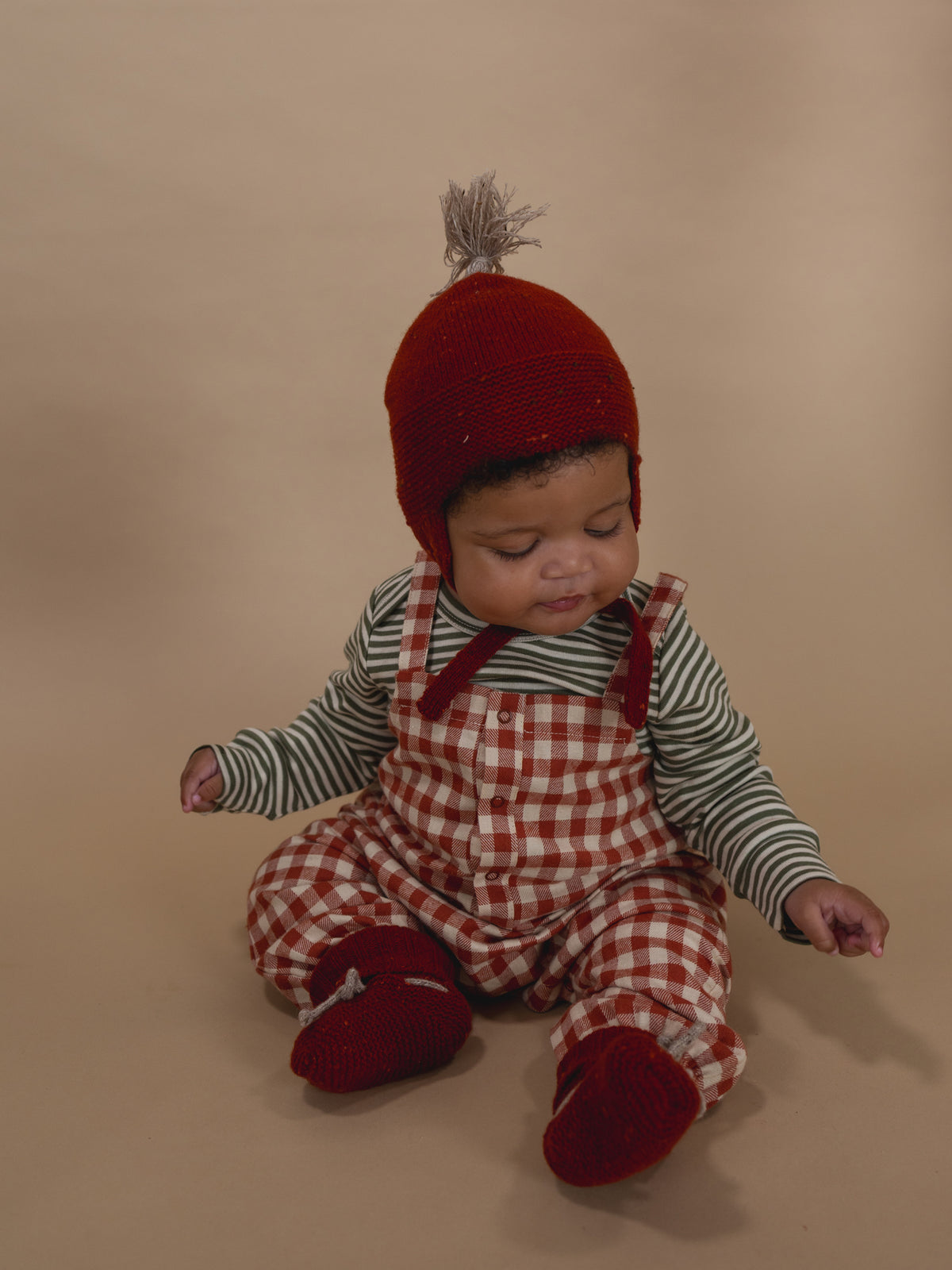 baby wearing paprika red wool tassel hat, matching booties, gingham dungarees and striped top