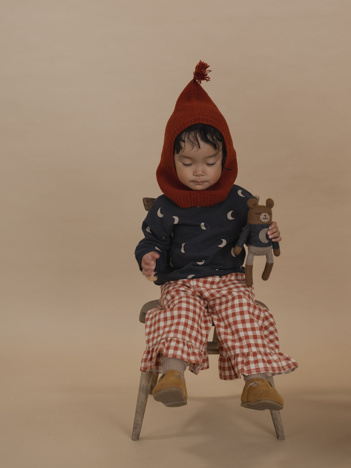 toddler in red wool balaclava with tassel, moon sweatshirt, gingham culottes, sitting on chair with toy