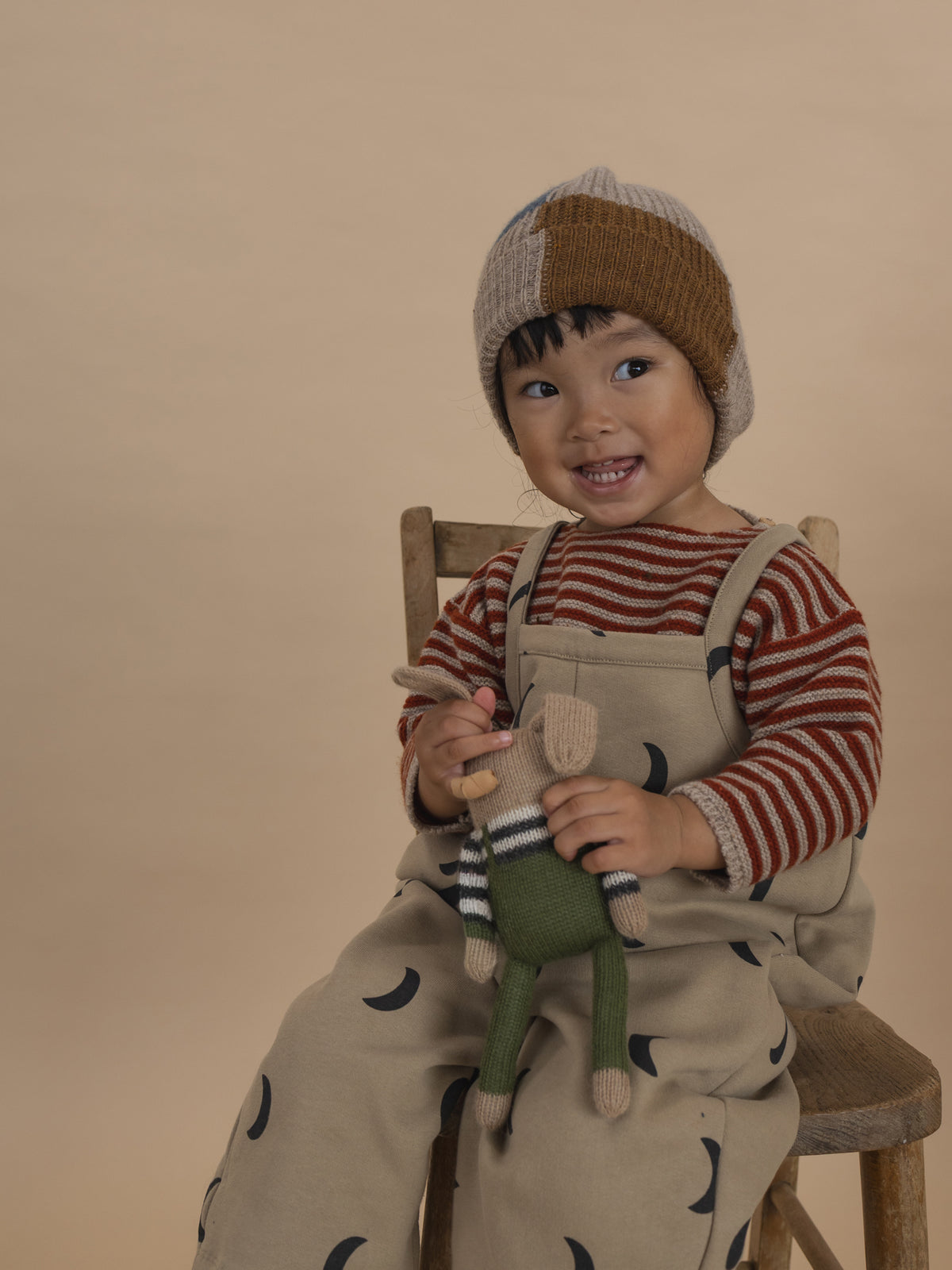 toddler in wool jumper with paprika red stripes, beige dungarees and patchwork beanie, holding plushie