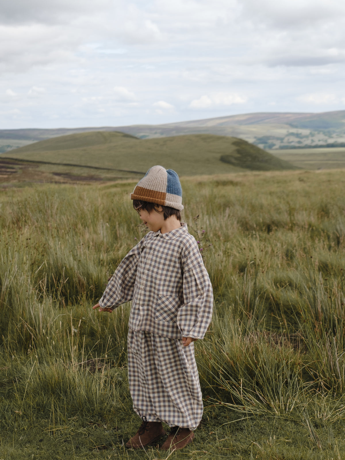 toddler in gingham lodge shirt with matching balloon pants & patchwork beanie, standing in grassy meadow