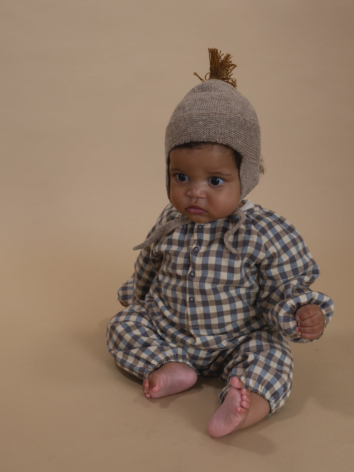 baby in cream and blue gingham flannel onesie, and oatmeal wool tassel hat sitting on floor