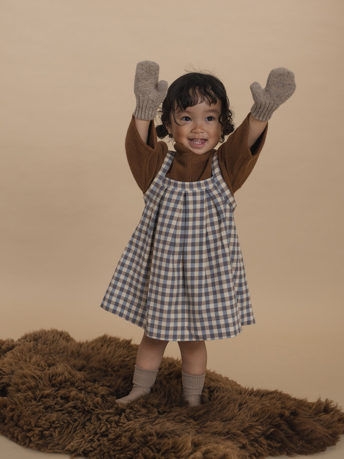 toddler in cream and blue gingham pleated pinafore skirt with jumper and mittens, standing with hands up