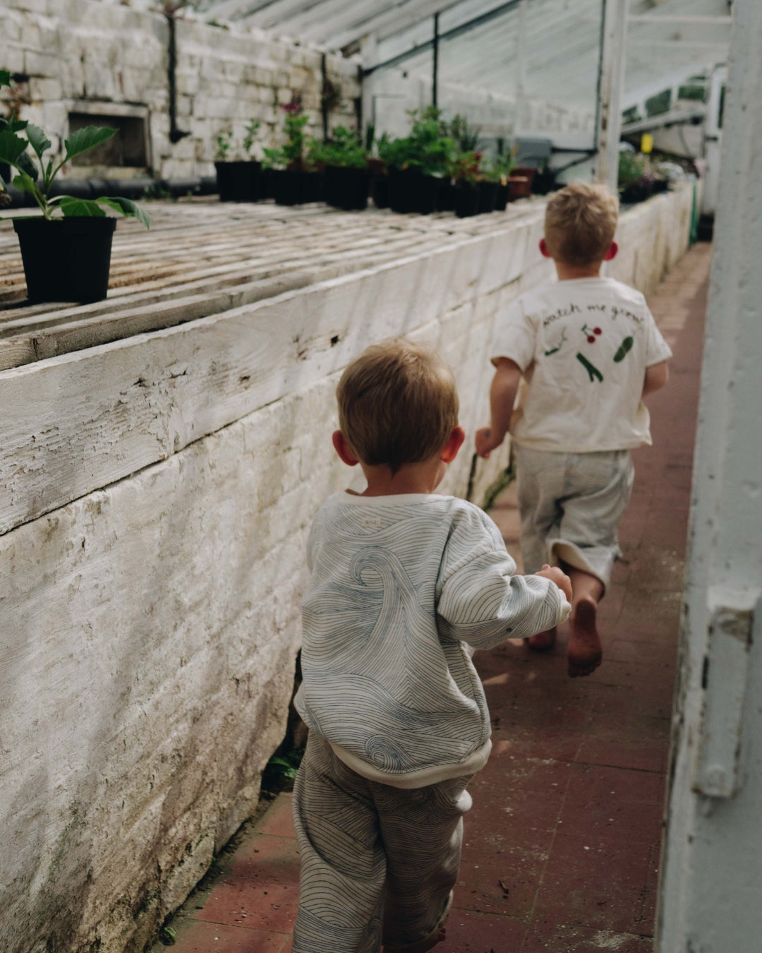 Two children walking through a greenhouse with plants and stone walls.
