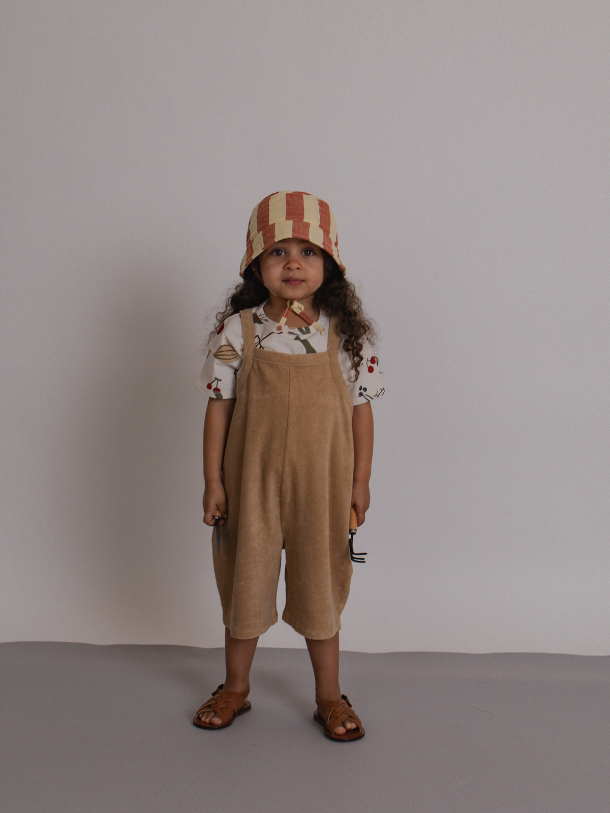 Child wearing a beige bucket hat and overalls against a plain background