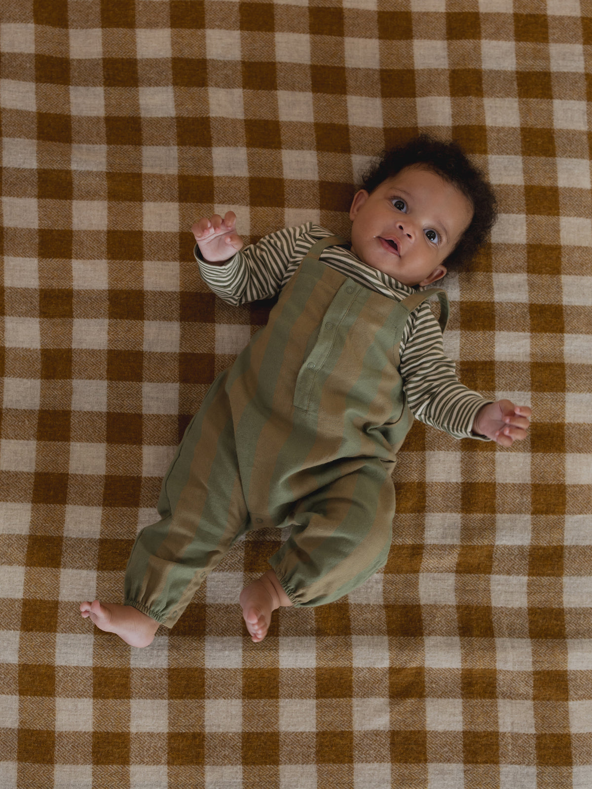 baby in green & cream gingham salopette and green striped sweatshirt, lying down on gingham blanket