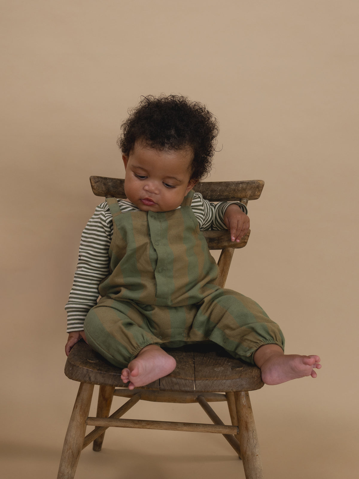 baby in green and cream gingham salopette with gathered cuffs, and striped sweatshirt sitting on chair