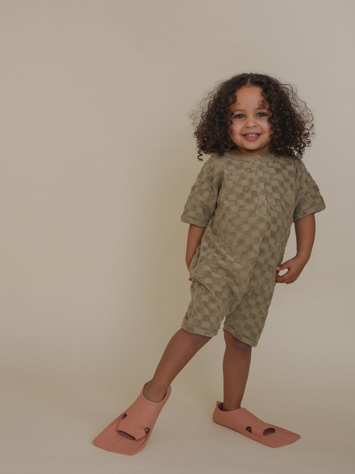 toddler in dusky beige checkerboard terry cotton beach romper standing on one leg with swimming fins
