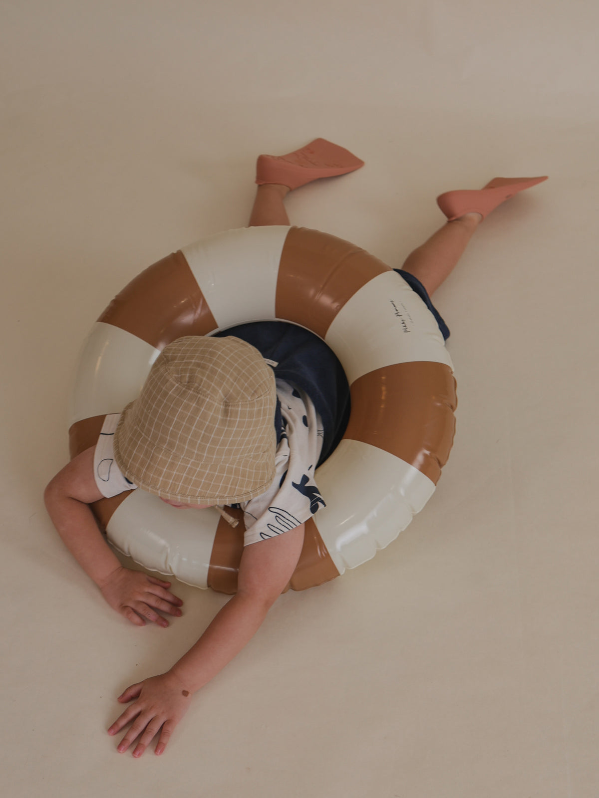 toddler in navy overalls, t-shirt, bucket hat & flippers, holding onto pool ring lying on floor pretending to swim
