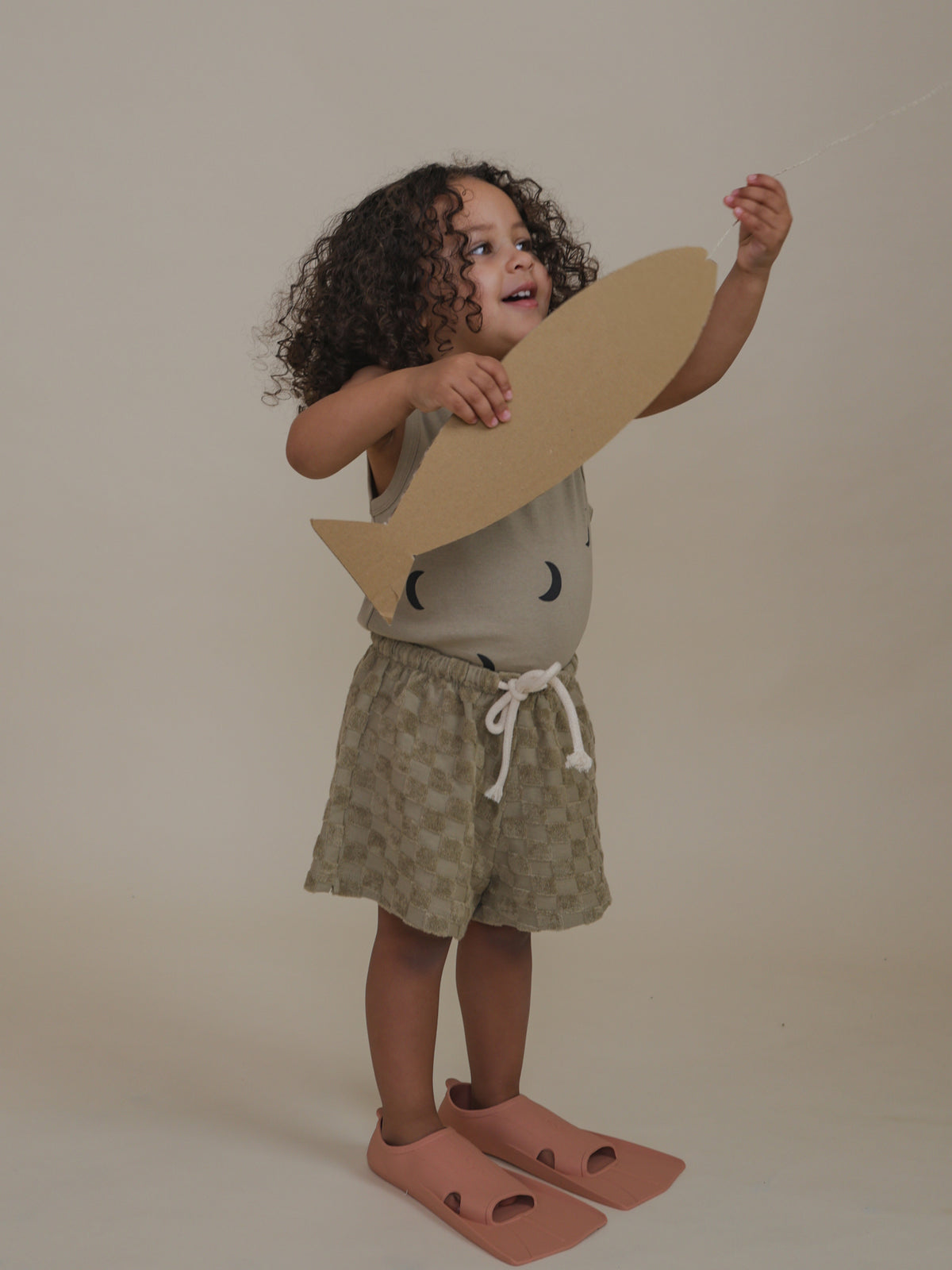toddler wearing beige checkerboard terry cotton rope shorts with swim fins holding cardboard fish