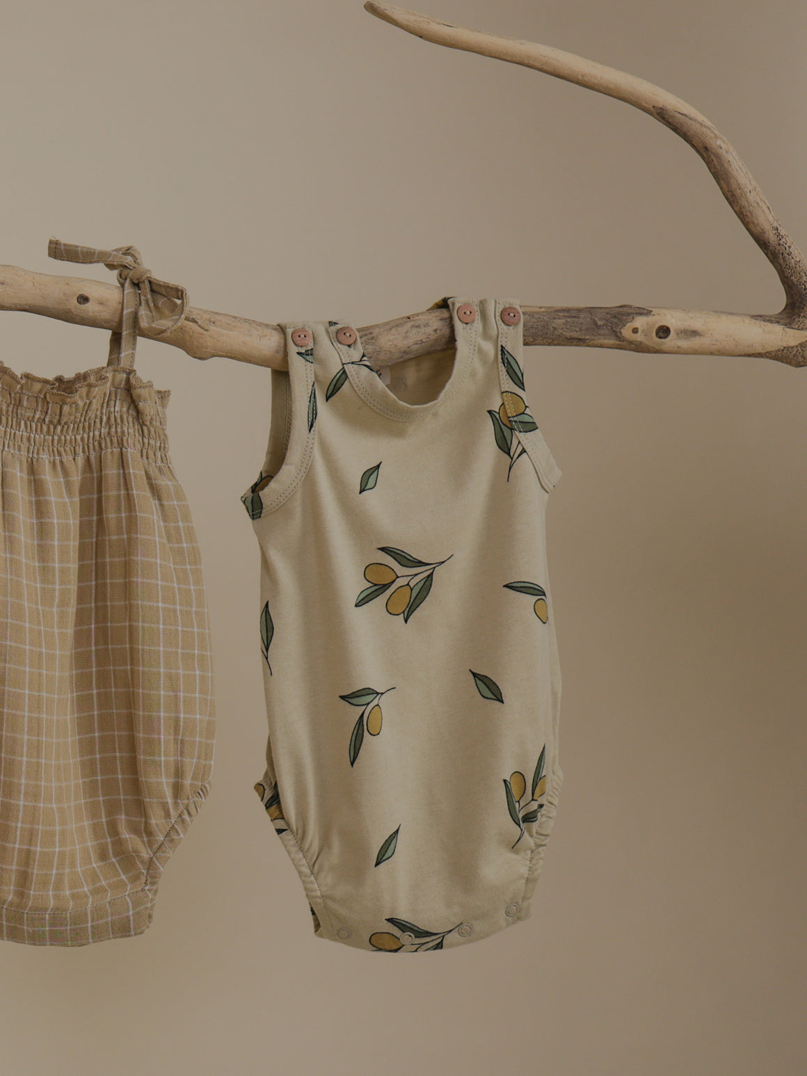 baby sleeveless cream bodysuit with olives & shoulder buttons, hanging on branch beside spaghetti bodysuit