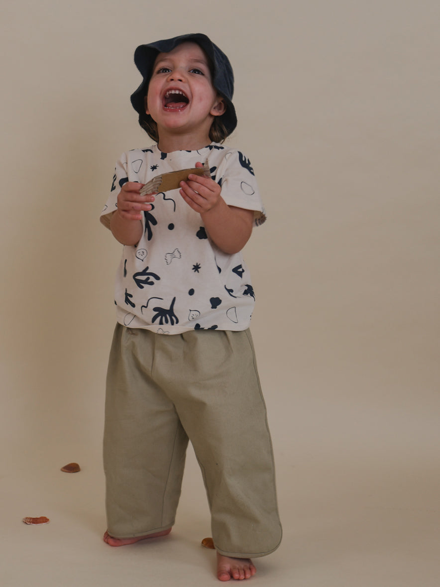 cheerful toddler wearing seasalt grey balloon trousers, printed t-shirt & sun hat, holding cardboard fish