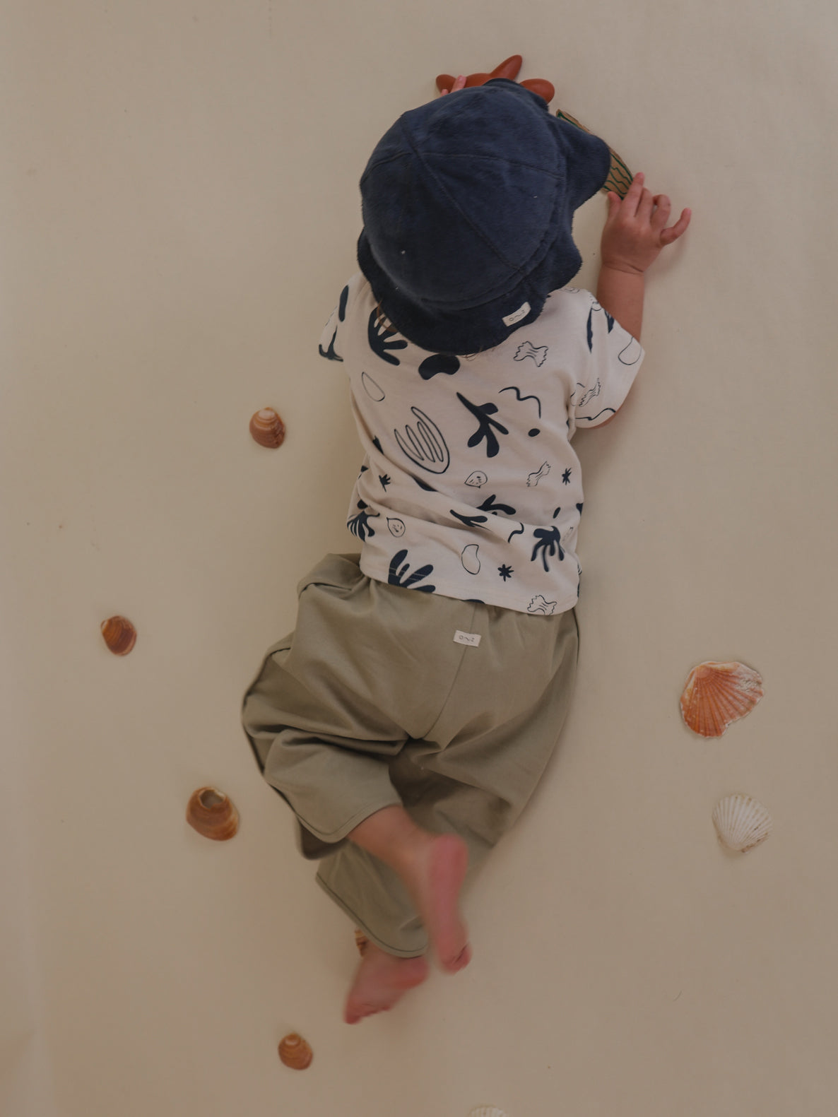toddler in navy terry sun hat, printed t-shirt & grey pants, playing on the floor with seashells