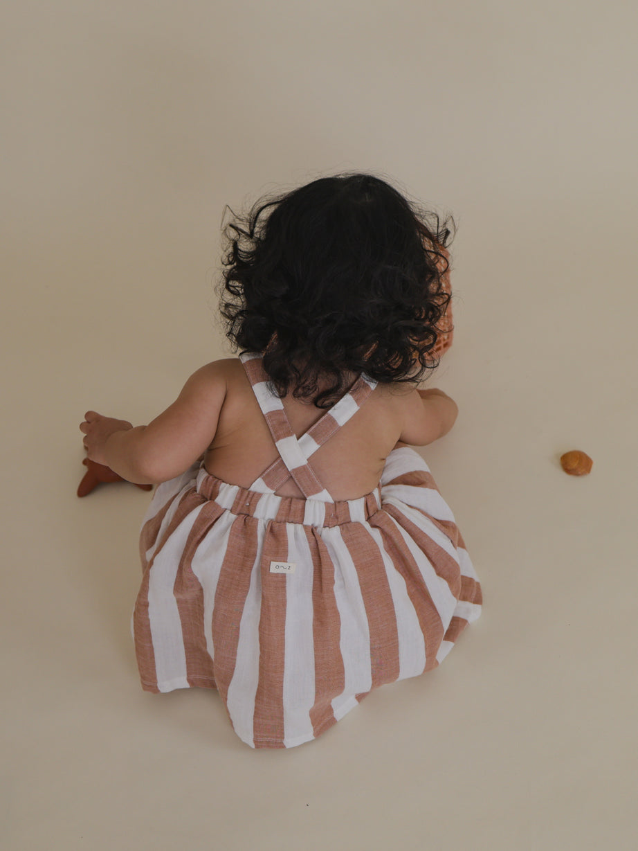 toddler in apron skirt with fun sienna red stripes & cross back crouching to pick seashells