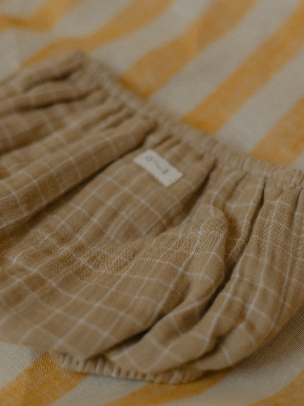 close up of soft breathable muslin baby shortie in wheat gold with grid pattern on striped mat