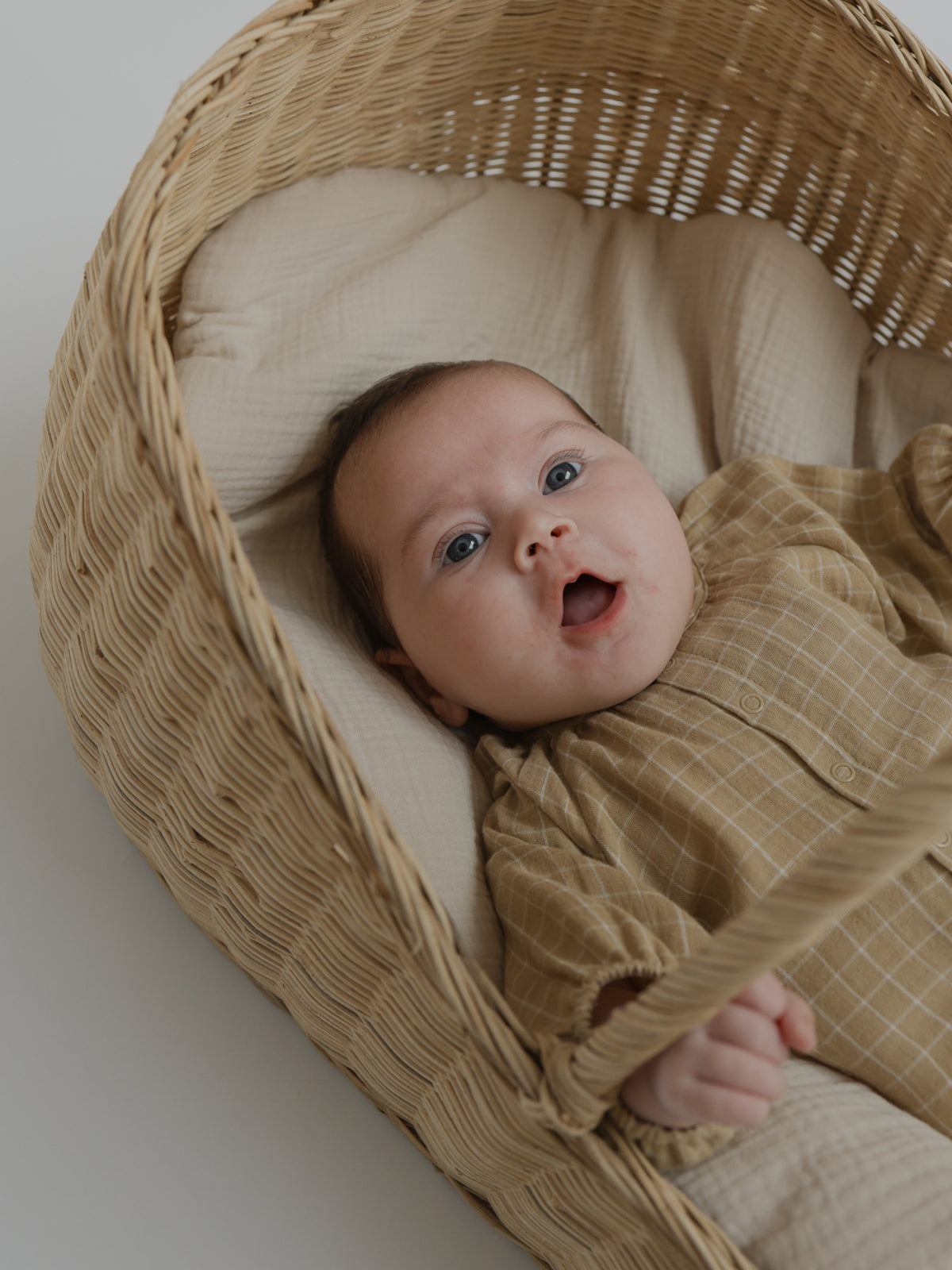 baby wearing muslin onesie in gold with grid pattern yawning in moses basket