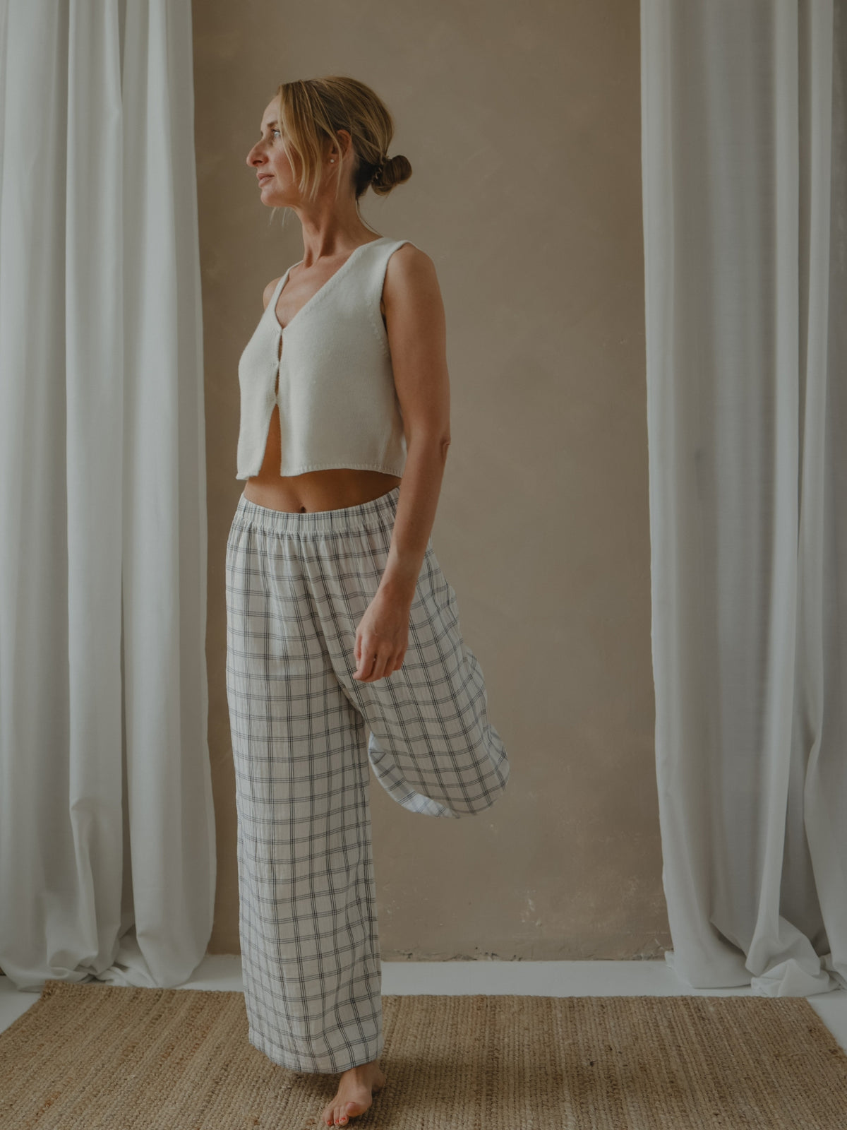 Woman wearing a white sleeveless top and checkered pants standing in front of white curtains.
