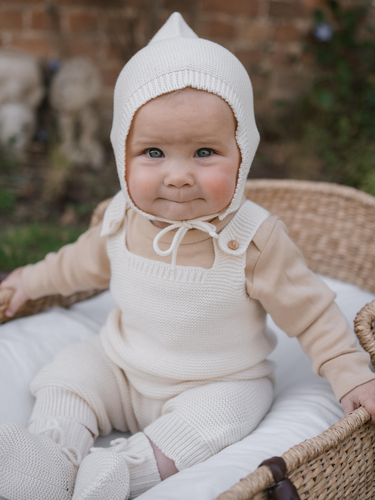happy baby in knitted cotton salopette and matching hat, booties, sitting in moses basket