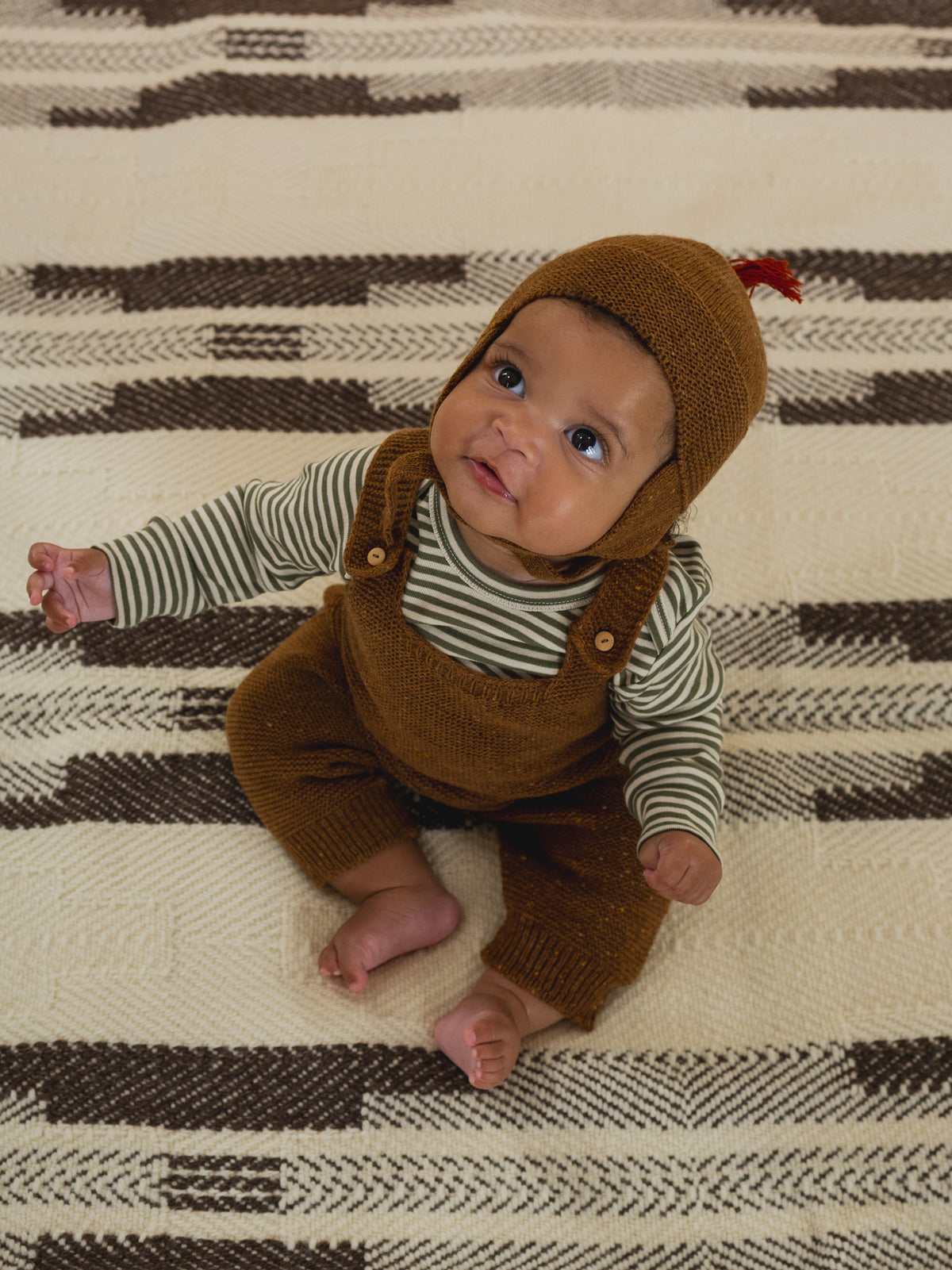 baby in chestnut brown wool hat with red tassel, matching wool salopette and striped top, looking up