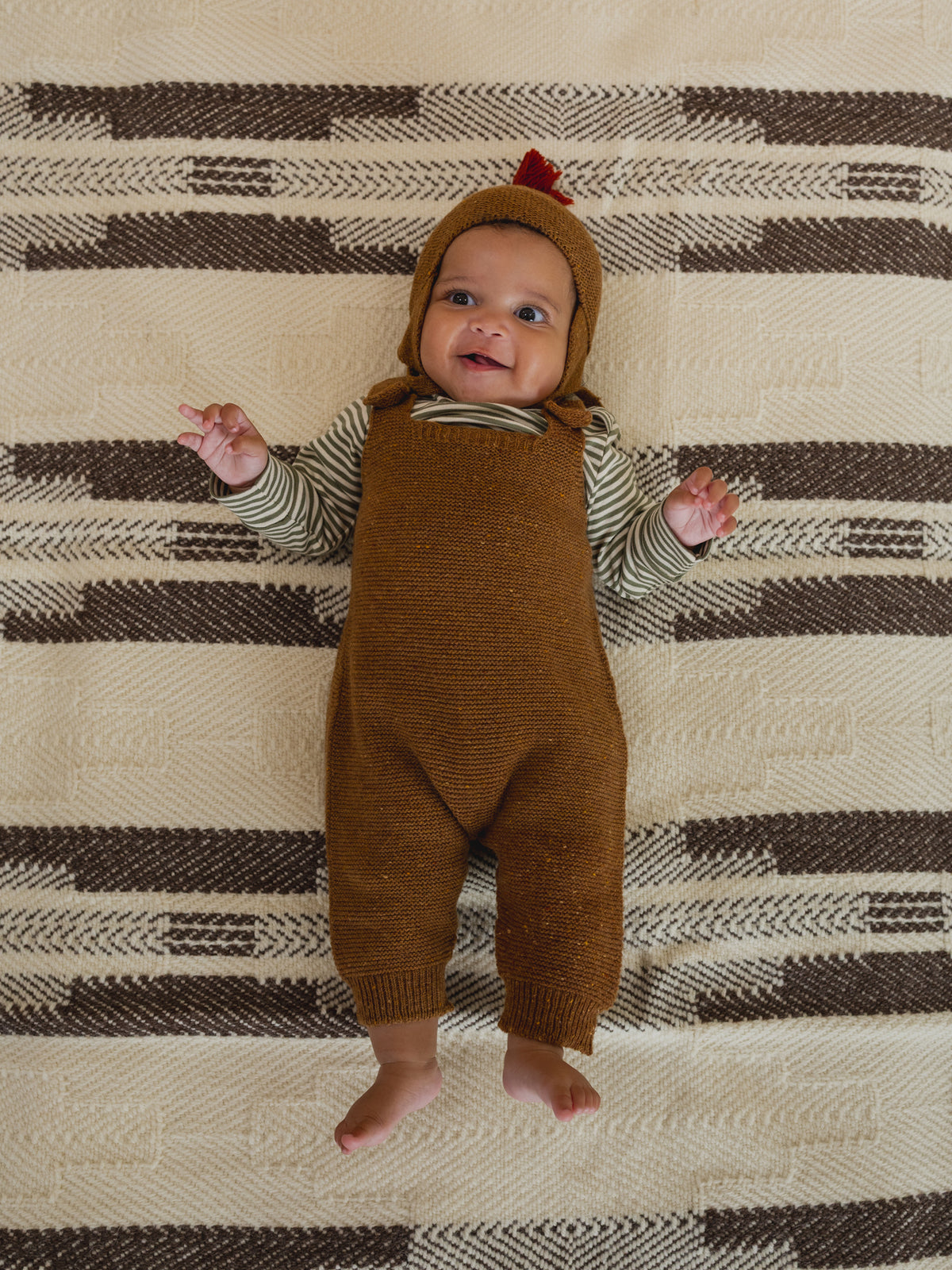 baby in chestnut brown wool hat with red tassel, matching wool salopette and striped top, lying down